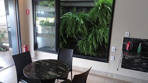 A room with a round table and chairs, a large window with a view of ferns, a fire extinguisher, and a wall-mounted electrical outlet. - Hotel Marina Torrano  (Trindade)