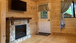 L’intérieur d’une cabane en rondins, avec une cheminée en pierre, un téléviseur fixé au mur et une porte avec des rideaux.