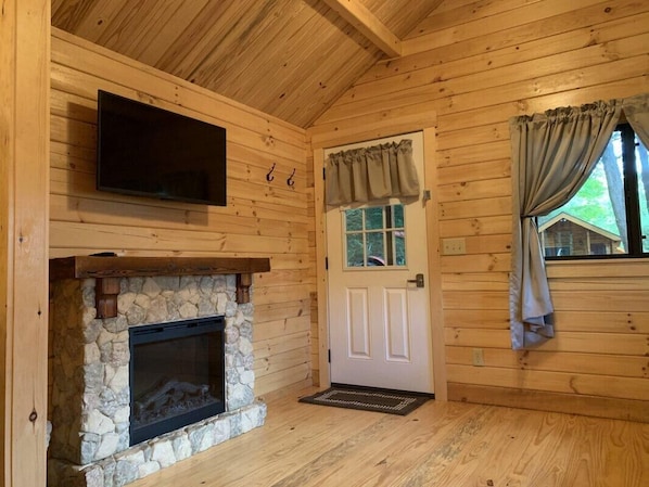 A log cabin interior with a stone fireplace, a television mounted on the wall, and a door with curtains.