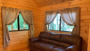 L’intérieur d’un chalet en bois, avec un canapé en cuir marron, deux fenêtres avec des rideaux et des murs en bois.