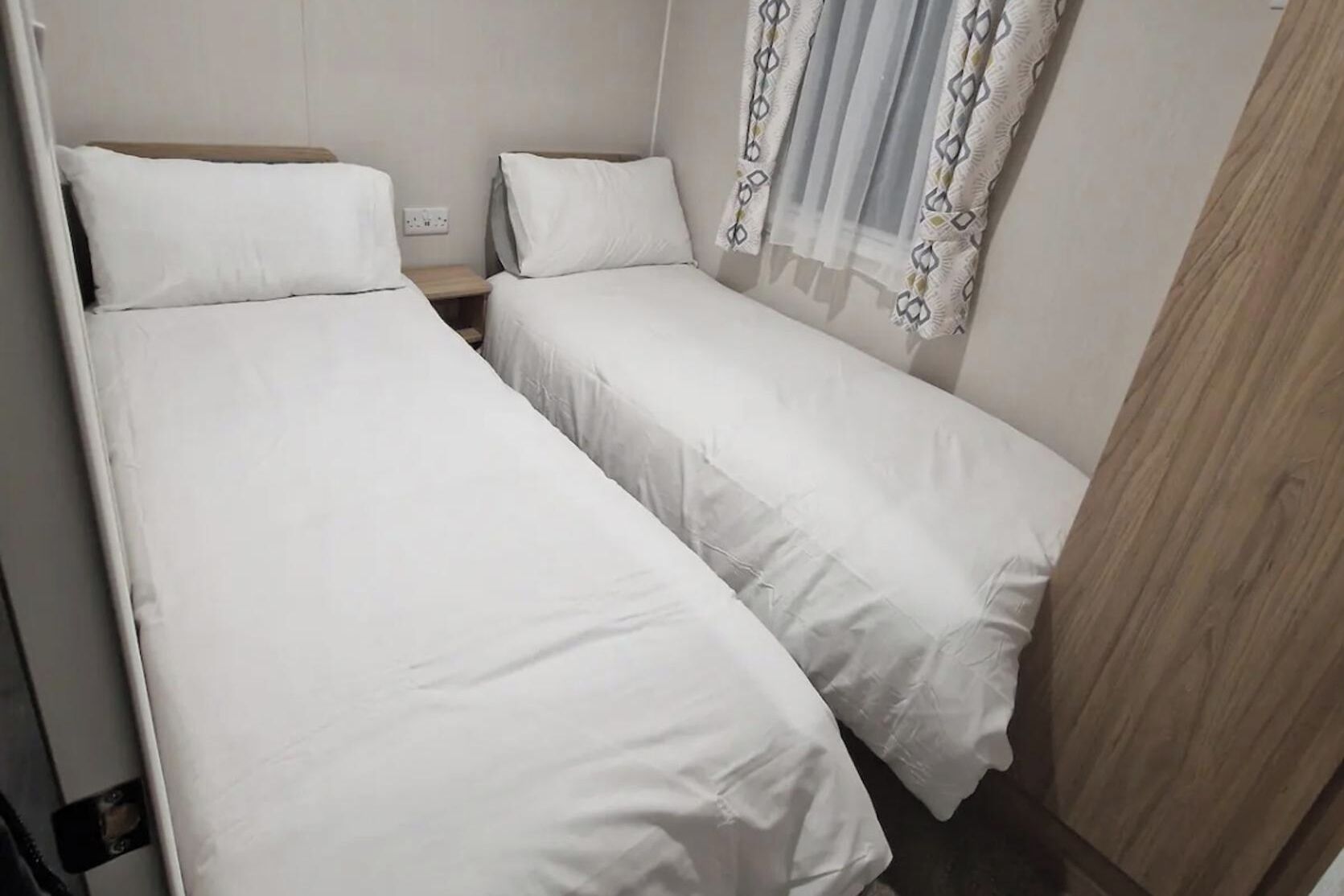 Une petite chambre propre, avec deux lits simples, une table de chevet en bois et une fenêtre agrémentée de rideaux à motifs.