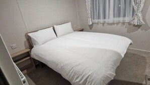 Un dormitorio con una cama individual, una mesita de noche, un enchufe y una ventana con cortinas translúcidas.