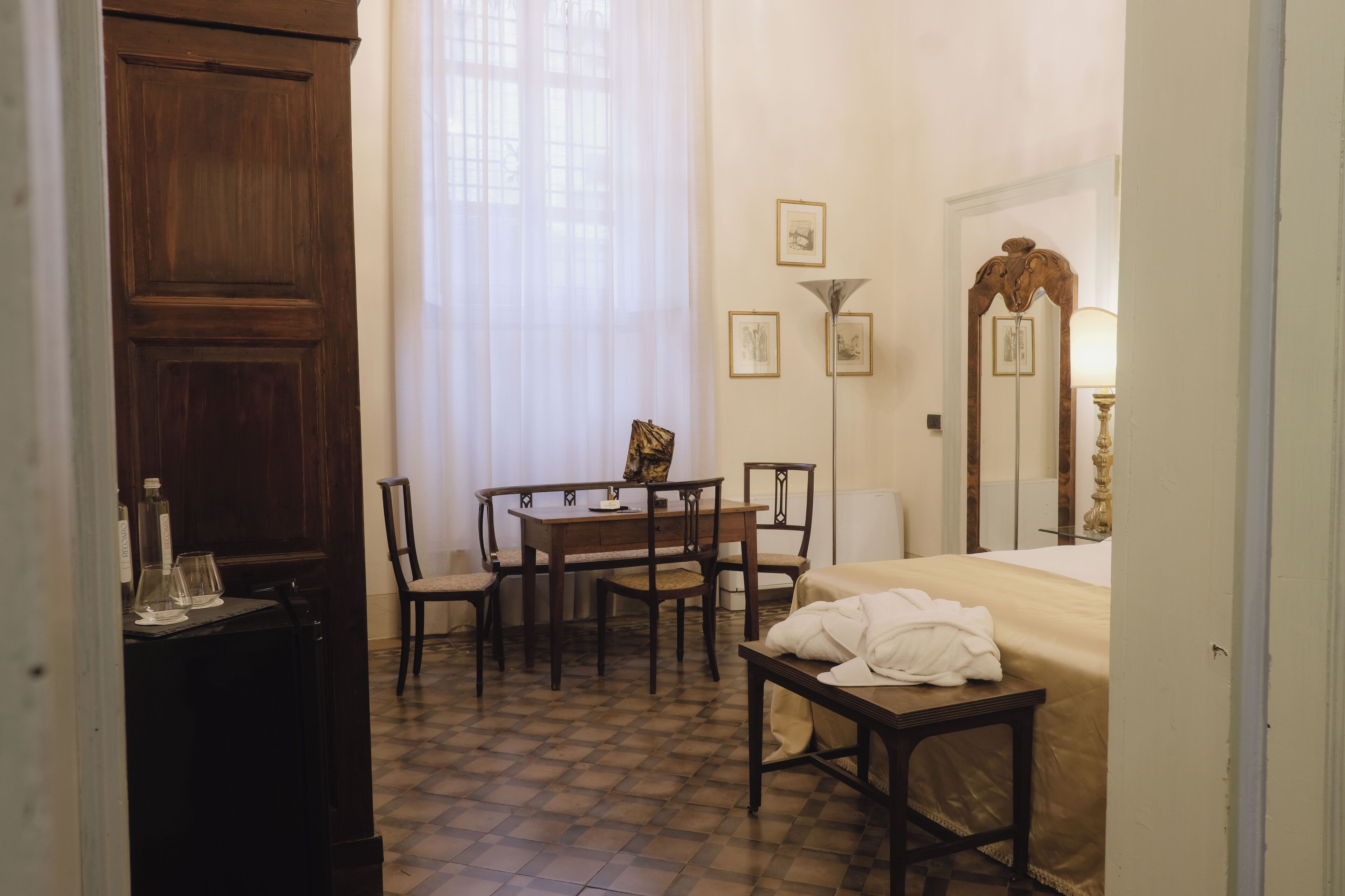Une chambre d’hôtel avec une armoire en bois, une table à manger avec des chaises, un lit avec une couette blanche et une lampe sur pied.