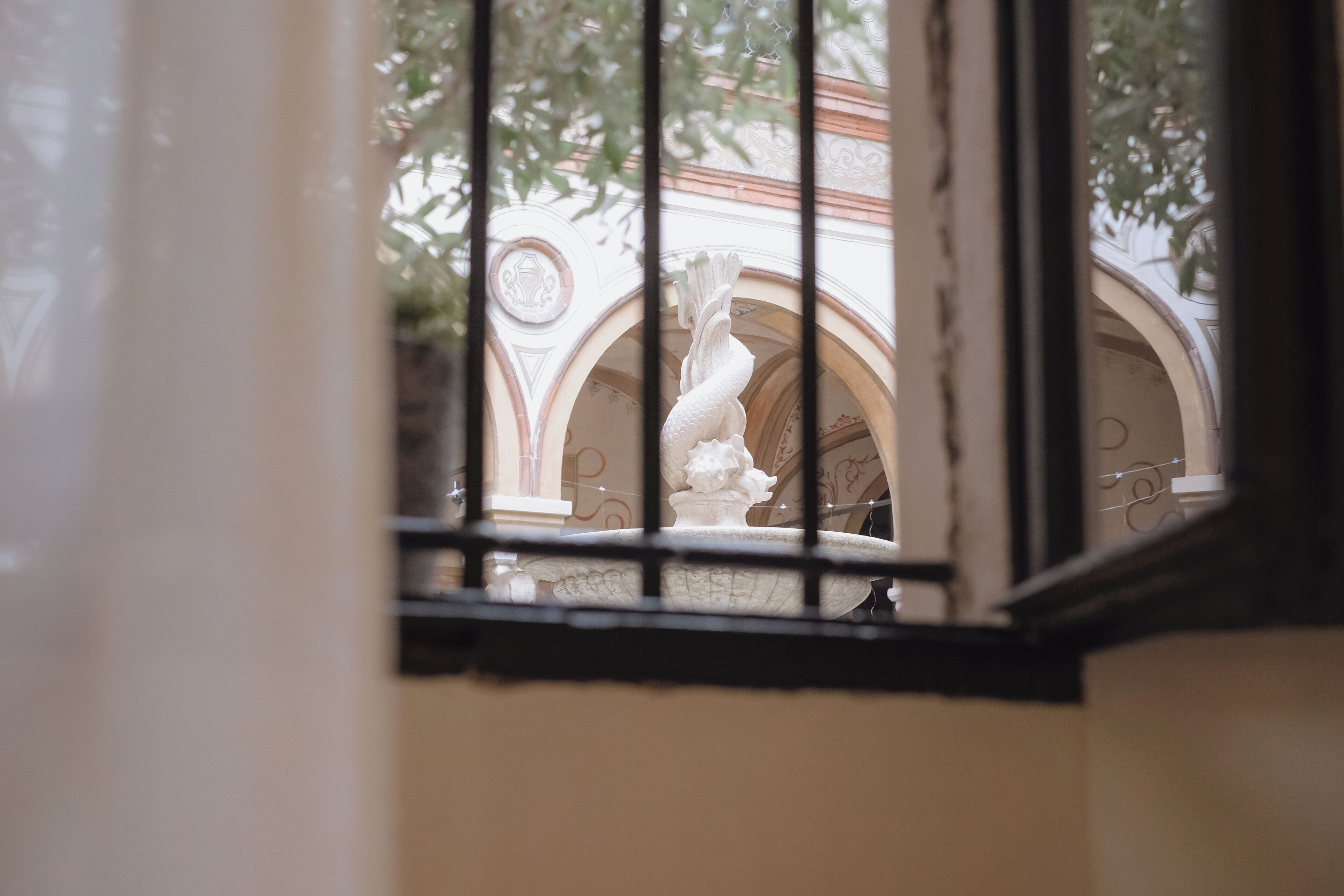 Une vue à travers une fenêtre, avec une statue blanche en arrière-plan, une balustrade en pierre et un portail en arc de cercle.