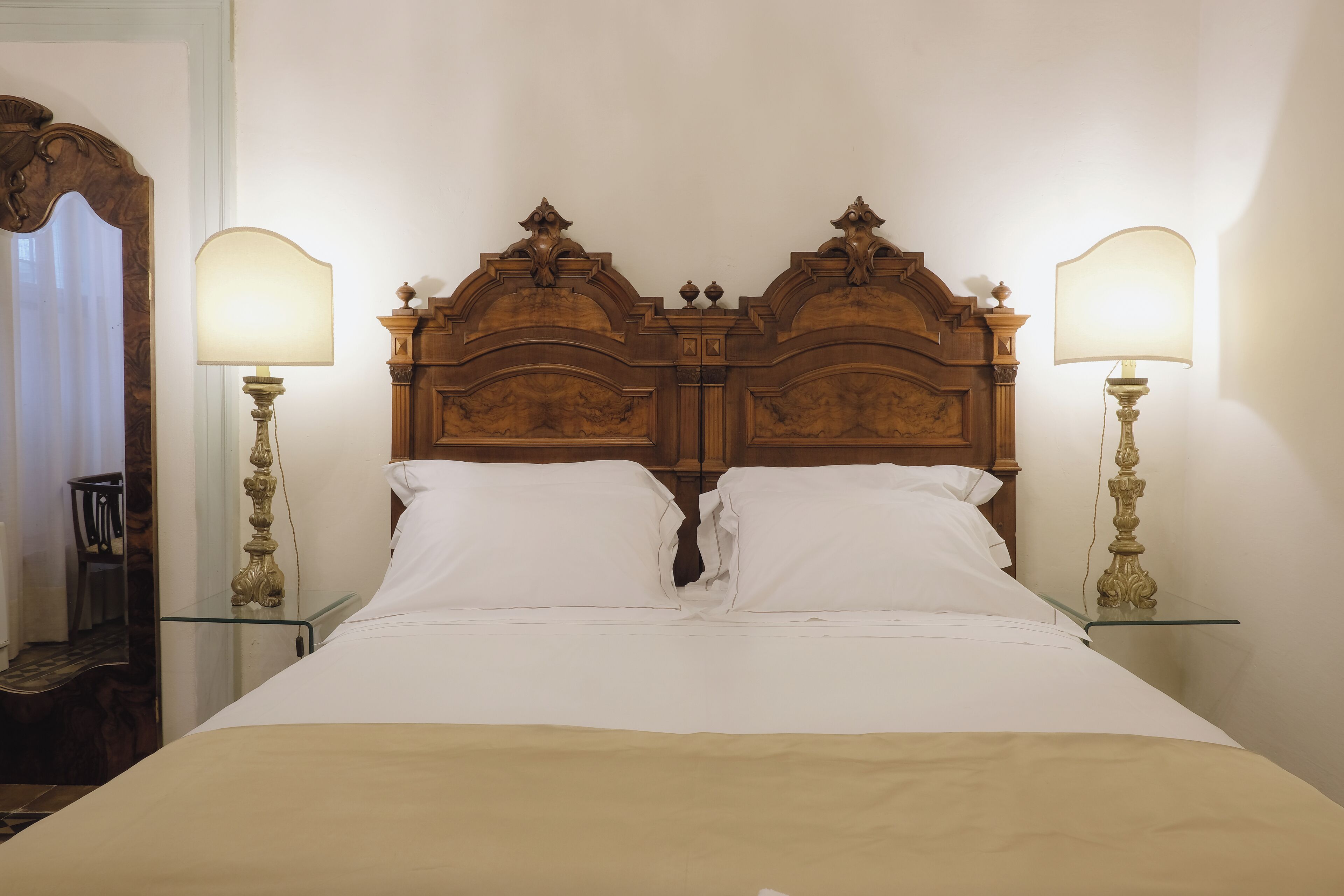 Una cama bien hecha con tablero de madera, dos lámparas de noche y una mesita auxiliar a su lado.