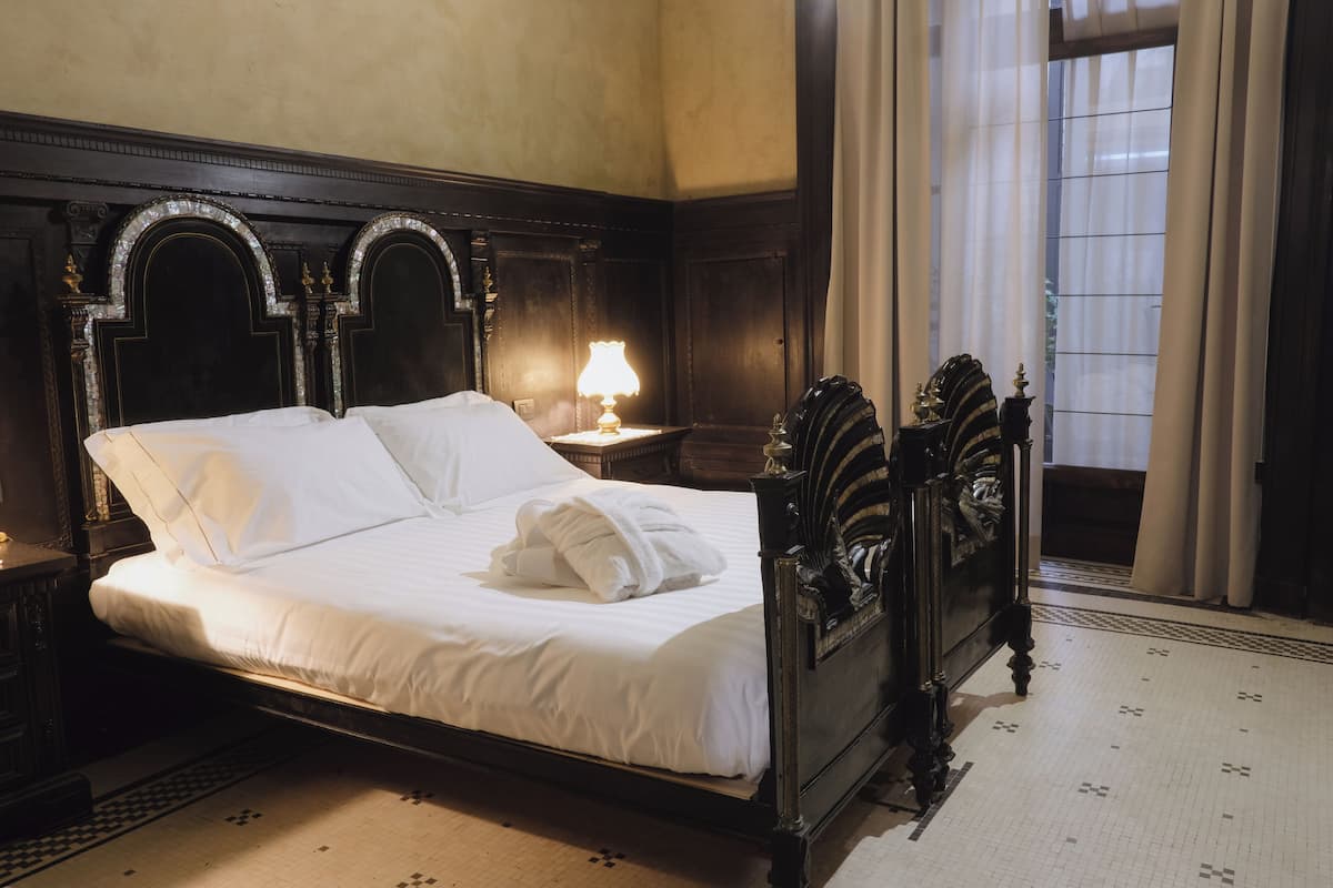 A neatly made bed with white linens and a folded towel, a bedside table with a lamp, and a dark wooden headboard with decorative elements.