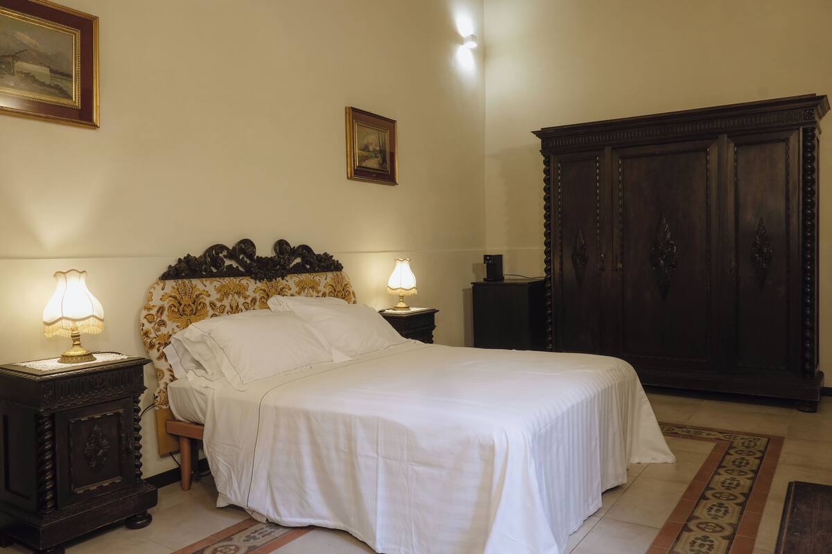 A neatly made bed with white linens, two bedside lamps, a wooden headboard, a dark wooden wardrobe, and a framed picture on the wall.