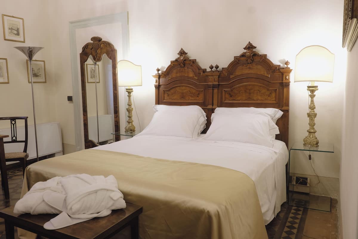 A hotel room with a large bed, two bedside lamps, a wooden headboard, a mirror, and a bedside table with a rolled-up towel.