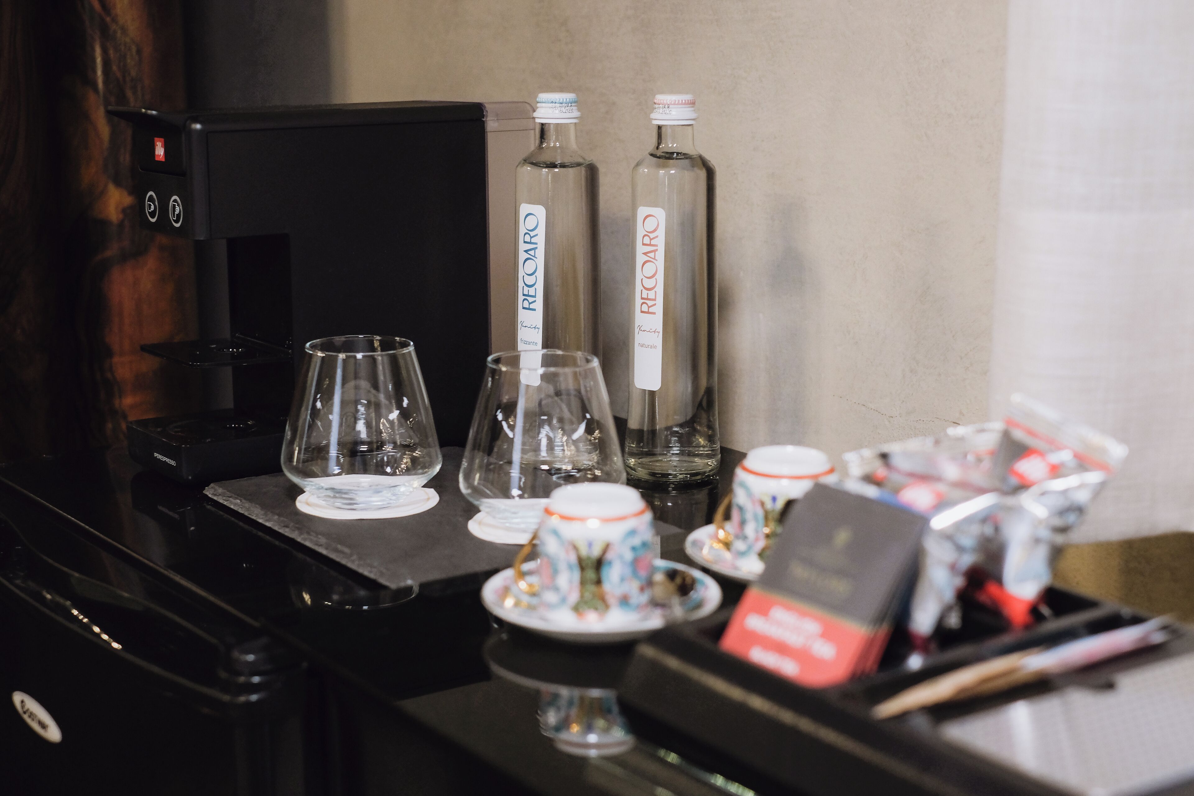 A coffee station with a coffee machine, two glass bottles labeled 'RECOARO', and several cups and saucers.