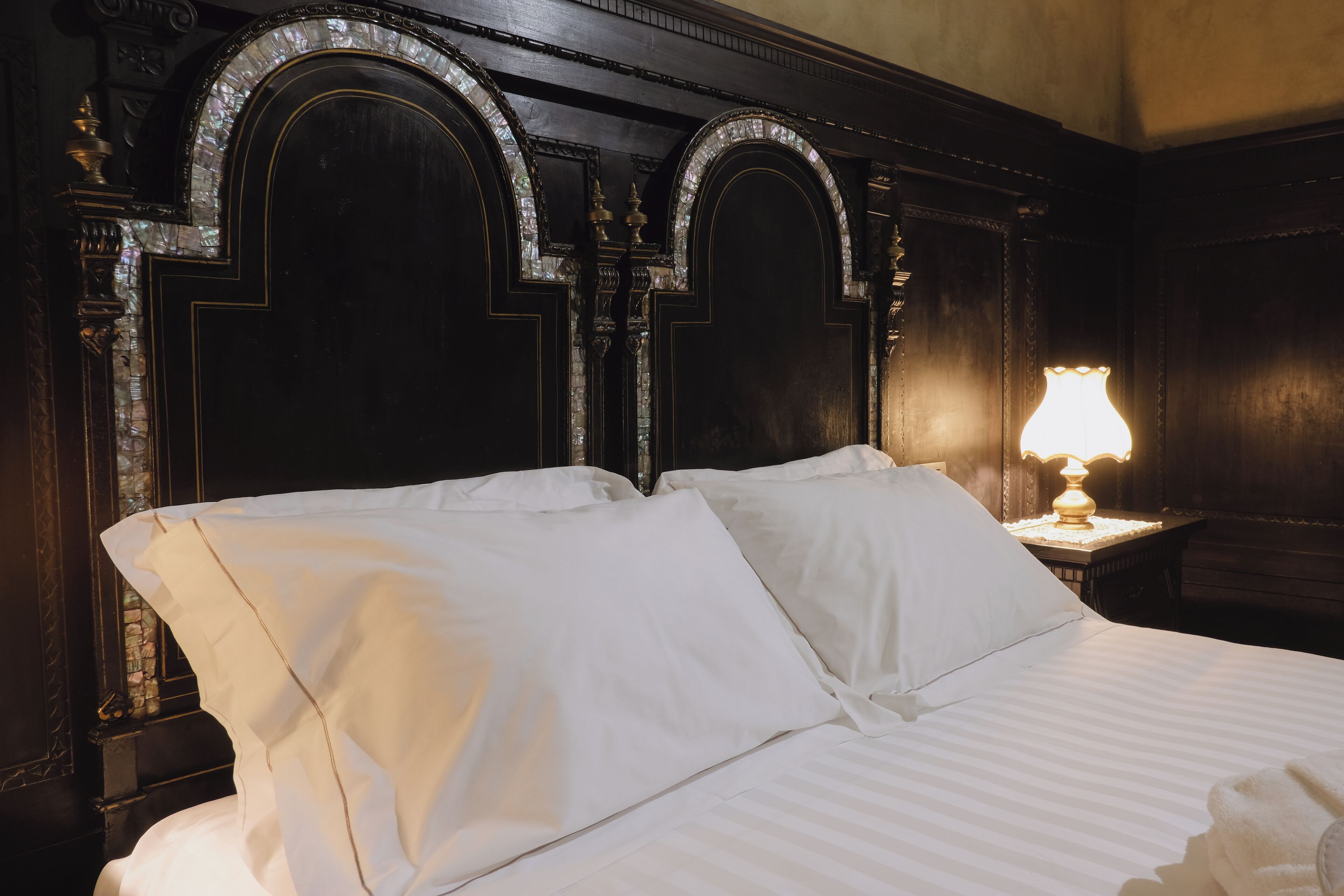 Una cama con sábanas blancas en una habitación con paneles de madera oscura y una lámpara encendida sobre una mesita de noche.