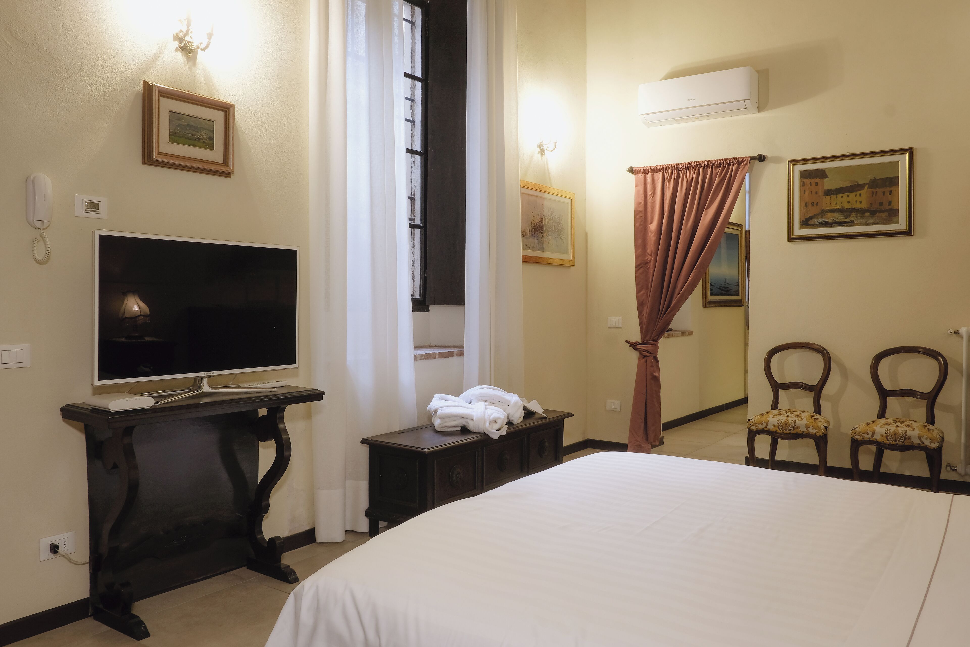 Une chambre d’hôtel avec un lit, un téléviseur posé sur un meuble, une petite table avec des serviettes, deux chaises et des œuvres d’art accrochées aux murs.