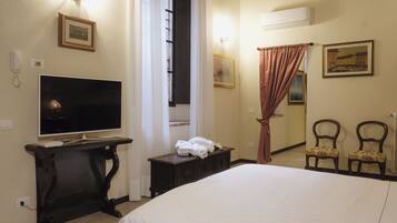 Une chambre d’hôtel avec un lit, un téléviseur posé sur un meuble, une petite table avec des serviettes, deux chaises et des œuvres d’art accrochées aux murs.
