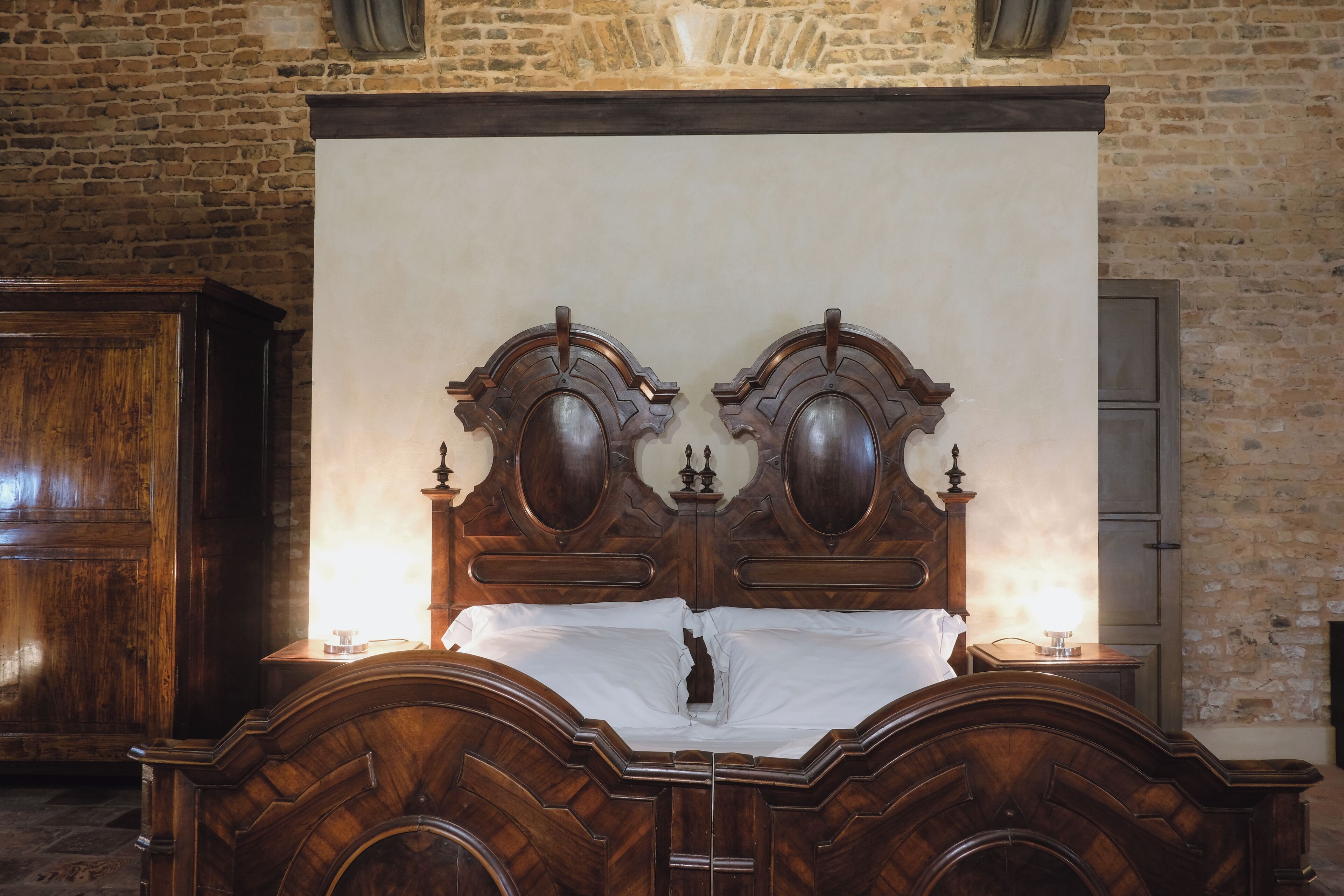 A wooden bed with a carved headboard, a nightstand, and a wall-mounted cabinet in a room with exposed brick walls.