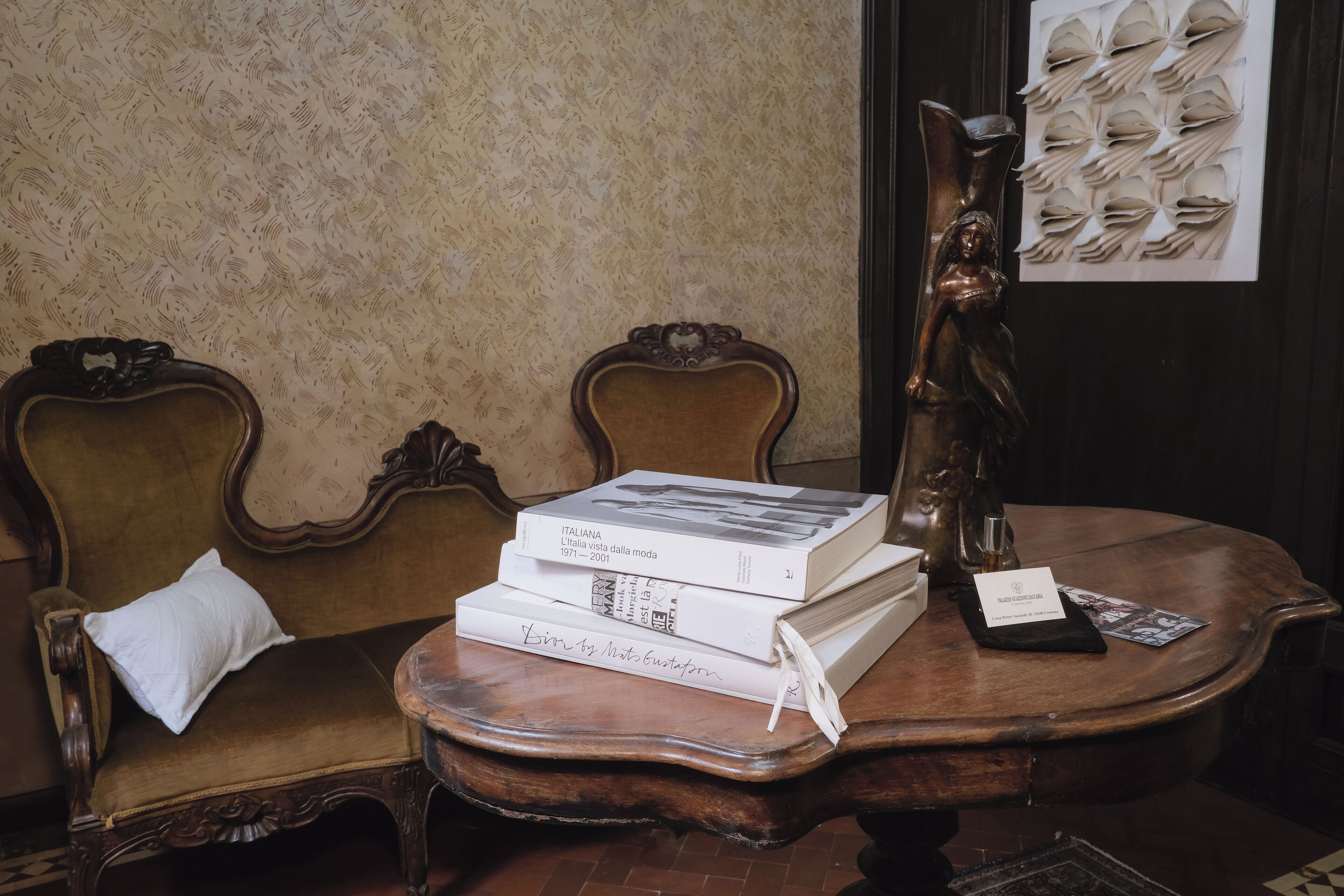 A room with a vintage wooden table, a stack of books, a white pillow on a chair, and a decorative angel statue.