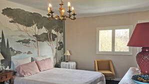 Een slaapkamer met een bed, een stoel, een tafel, een lamp, een kroonluchter, een raam en een muurschildering.