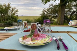 A picnic setup with a plate of food, utensils, and a glass pitcher on a table with a view of a field and trees. - Entre Mont St Michel et Merveille (Vains)