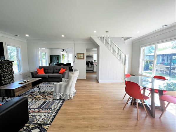 A spacious living room with a sofa, a dining table with chairs, a staircase, and a kitchen in the background. - Tranquil Kingsville Cottage - Private Beach & Pet-Friendly! (Kingsville)