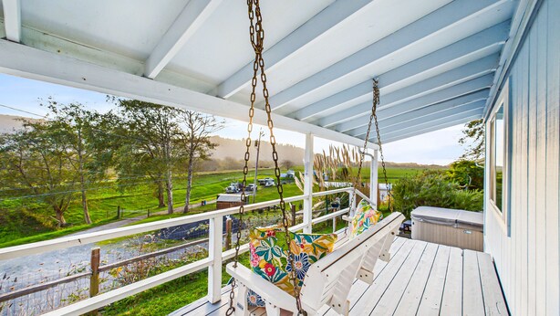 A porch with a swing, a view of a river, and a grassy area with trees. - Explore Redwood National Park with Hot Tub, Wood Stove, and Fire Pit! (Orick)