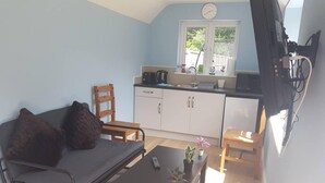 A compact living space with a sofa, a small table with a remote control, a chair, a kitchen area with cabinets and a window, and a clock on the wall. - Cozy Studio in Luton (Luton)