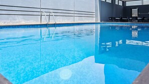 En klar swimmingpool med en metalstige, omgivet af en fliseterrasse og en væg med vandrette linjer.