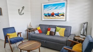 En stue med en grå sofa, blå lænestole og et træbord. Der er et maleri af både på væggen og en lille sejlbåd på et sofabord.