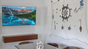 Et spiseområde med et glasbord, træstole med blå hynder, et ophængt tv der viser et bjerglandskab, og en dekorativ vægophængning med maritimt tema.