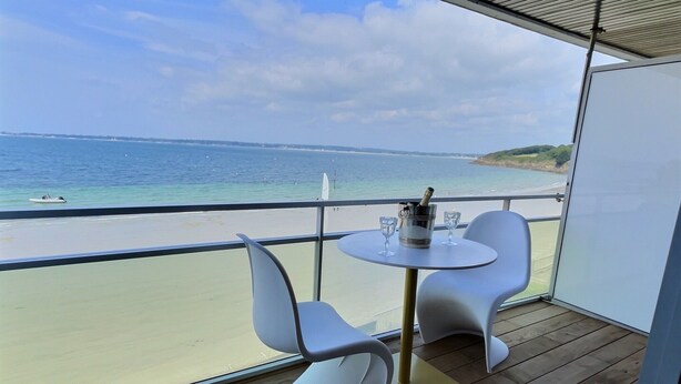 Un balcon doté d’un plancher en bois, de deux chaises blanches, d’une petite table ronde avec une bouteille et des verres, et offrant une vue sur la plage et l’océan.