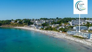 Vue aérienne d’une plage aux eaux turquoise et limpides, bordée d’une étendue de sable et d’une rangée de bâtiments le long de la côte.