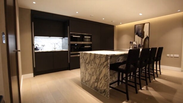 A modern kitchen with dark cabinetry, a marble island, and bar stools. - Luxurious Three Bedroom Flat in Chelsea Creek (London)