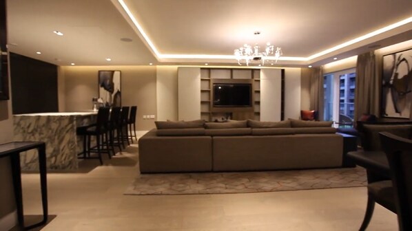 A modern living room with a large sectional sofa, a dining area with chairs, a TV, and a chandelier. - Luxurious Three Bedroom Flat in Chelsea Creek (London)