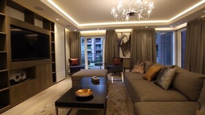 A modern living room with a large flat-screen TV, a sofa with multiple cushions, a coffee table with a bowl, and a chandelier. - Luxurious Three Bedroom Flat in Chelsea Creek (London)