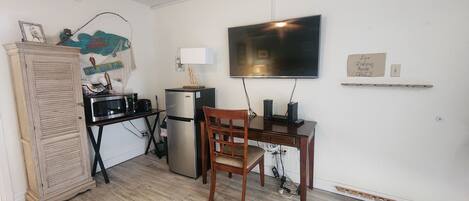 A compact kitchen with a refrigerator, microwave, and a dining table with a chair. A television is mounted on the wall above the table.