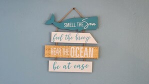 A wall decoration with a whale and phrases like "SMELL THE SEA," "feel the breeze," "HEAR THE OCEAN," and "be at ease." - Spring Pricing at Seaside Bungalow (Rockport)