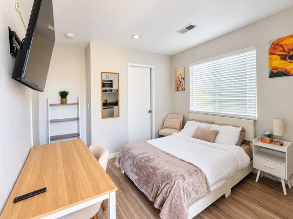 A bedroom with a bed, bedside table, chair, TV mounted on the wall, and a small kitchenette.