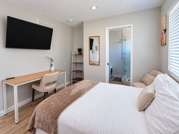 A modern bedroom with a bed, a desk, a chair, a TV, and a walk-in shower.