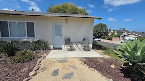 A single-story house with a front door, two chairs, and a small table on a concrete patio.