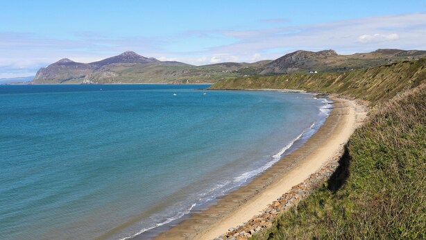 A coastal landscape with a sandy beach, clear blue waters, and distant mountains. - Maes Y Ddol (Pwllheli)