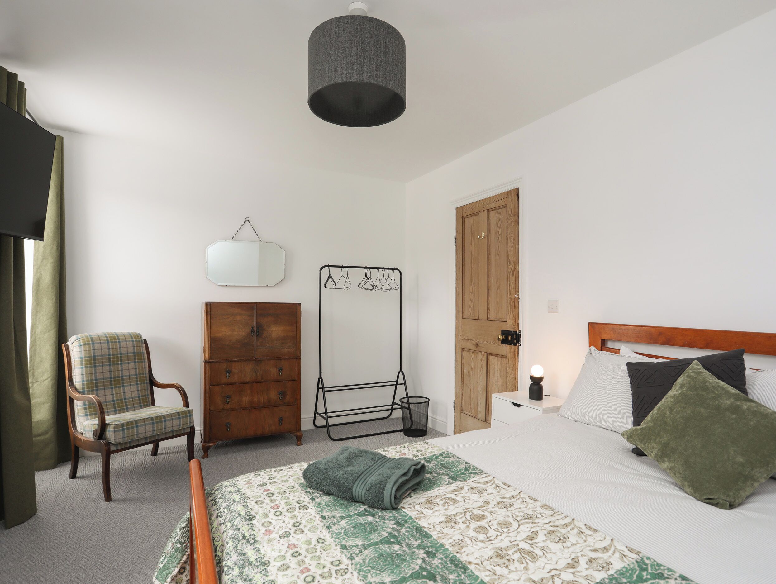 Une chambre à coucher comprenant un lit, une chaise, une commode en bois, un porte-manteau et une porte.