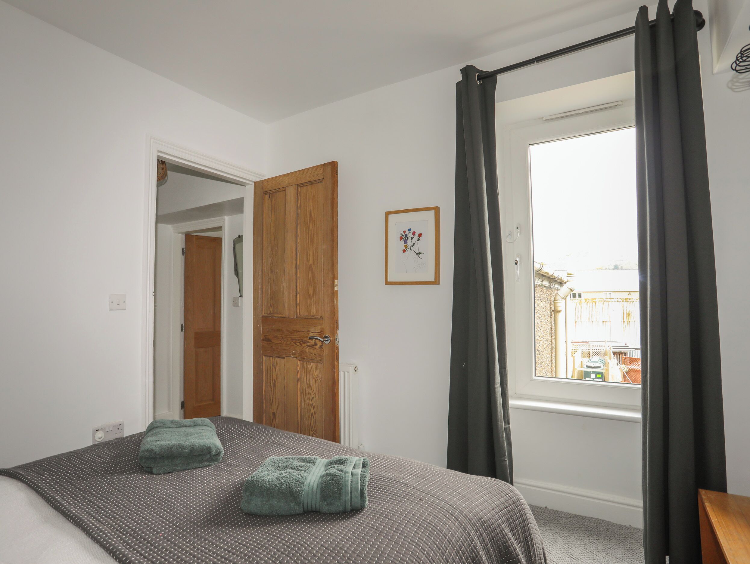 A bedroom with a bed, a wooden door, a window with curtains, and a small framed picture on the wall.
