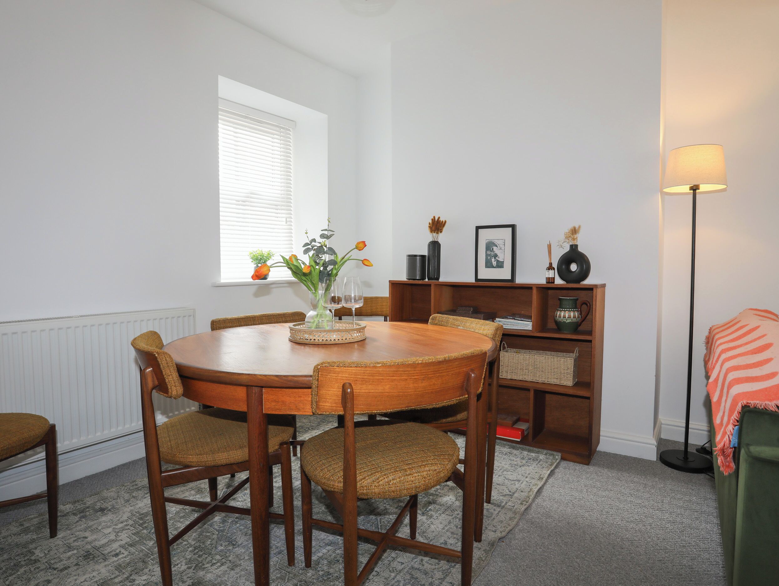 Een eetruimte met een houten tafel en stoelen, een vaas met bloemen en een boekenplank met diverse objecten.