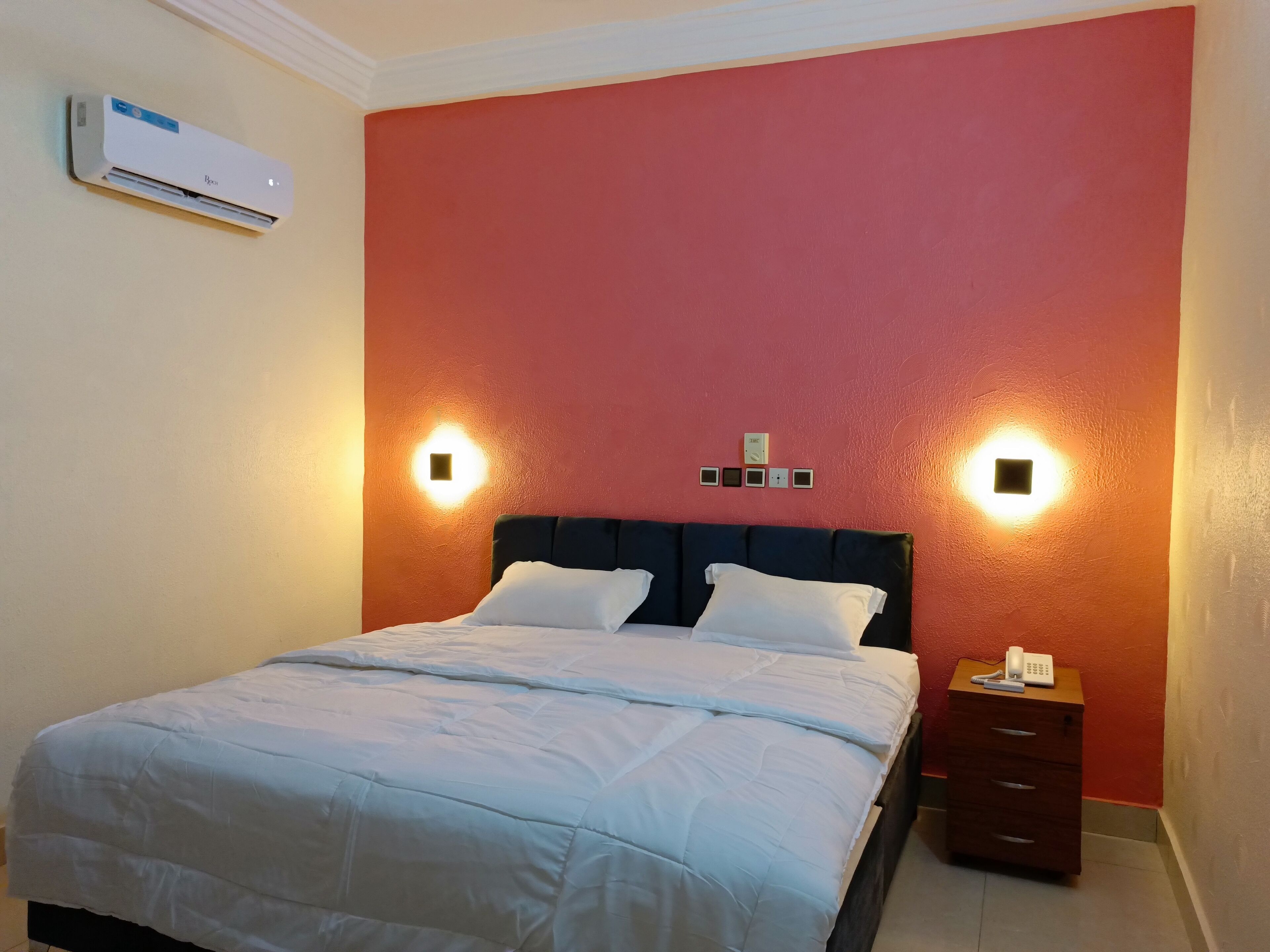 A neatly made bed with a white comforter and pillows, a dark headboard, and a bedside table with a phone and remote control.