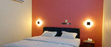 A neatly made bed with a white comforter and pillows, a dark headboard, and a bedside table with a phone and remote control.
