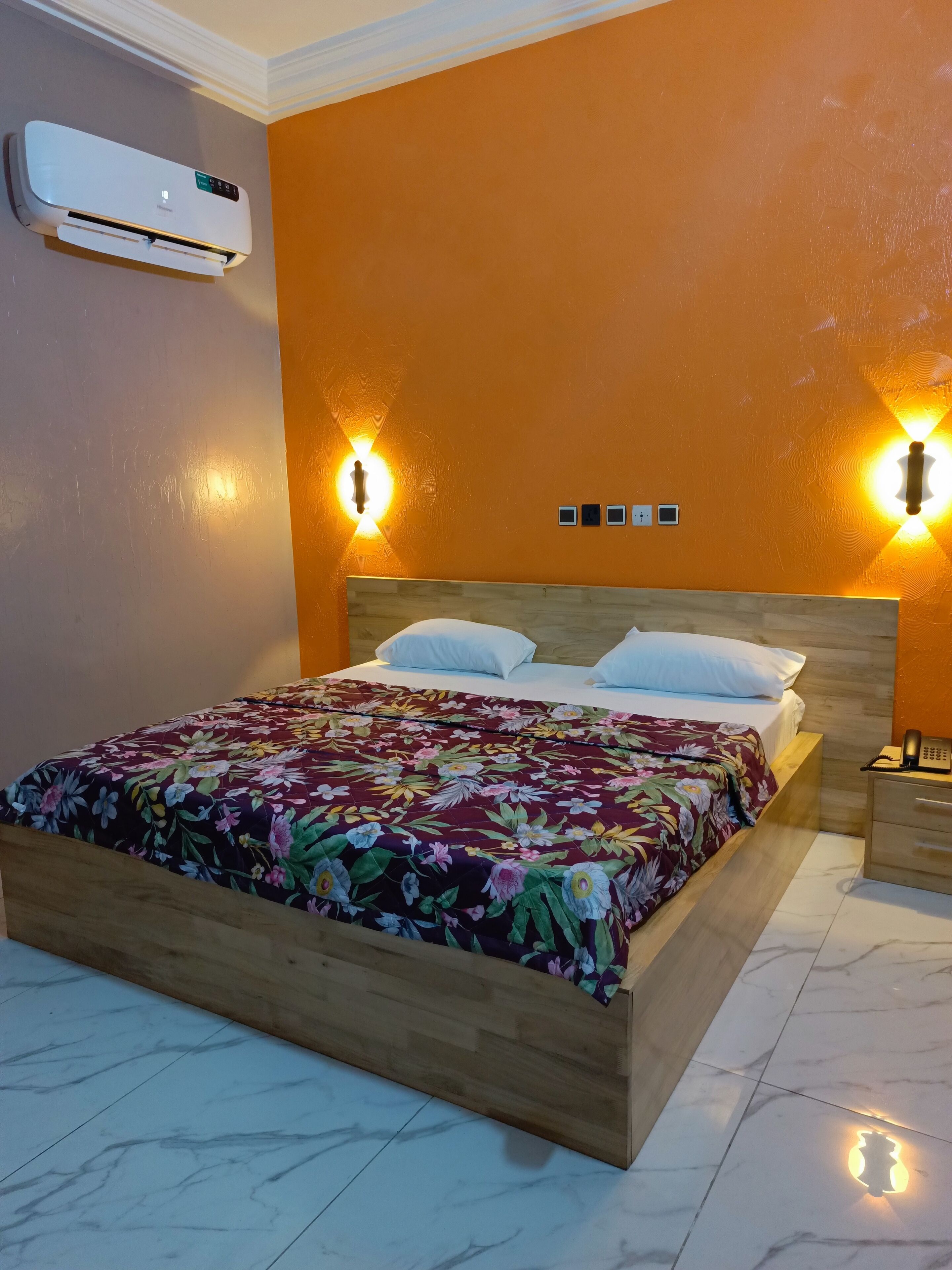 Une chambre avec un lit en bois, une literie à motifs floraux, un climatiseur et des lampes fixées au mur.