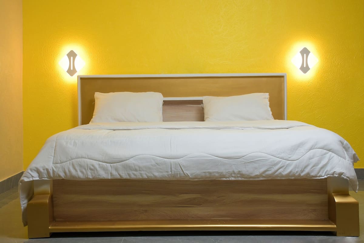 Un lit avec un cadre en bois et une literie blanche, placé devant un mur jaune orné de deux appliques murales.