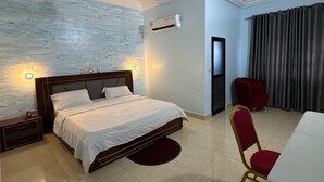 A hotel room with a bed, bedside tables, a desk, a chair, an air conditioning unit, a ceiling fan, and a window with curtains. - HOTEL MAVILLA Cotonou (Cotonou)