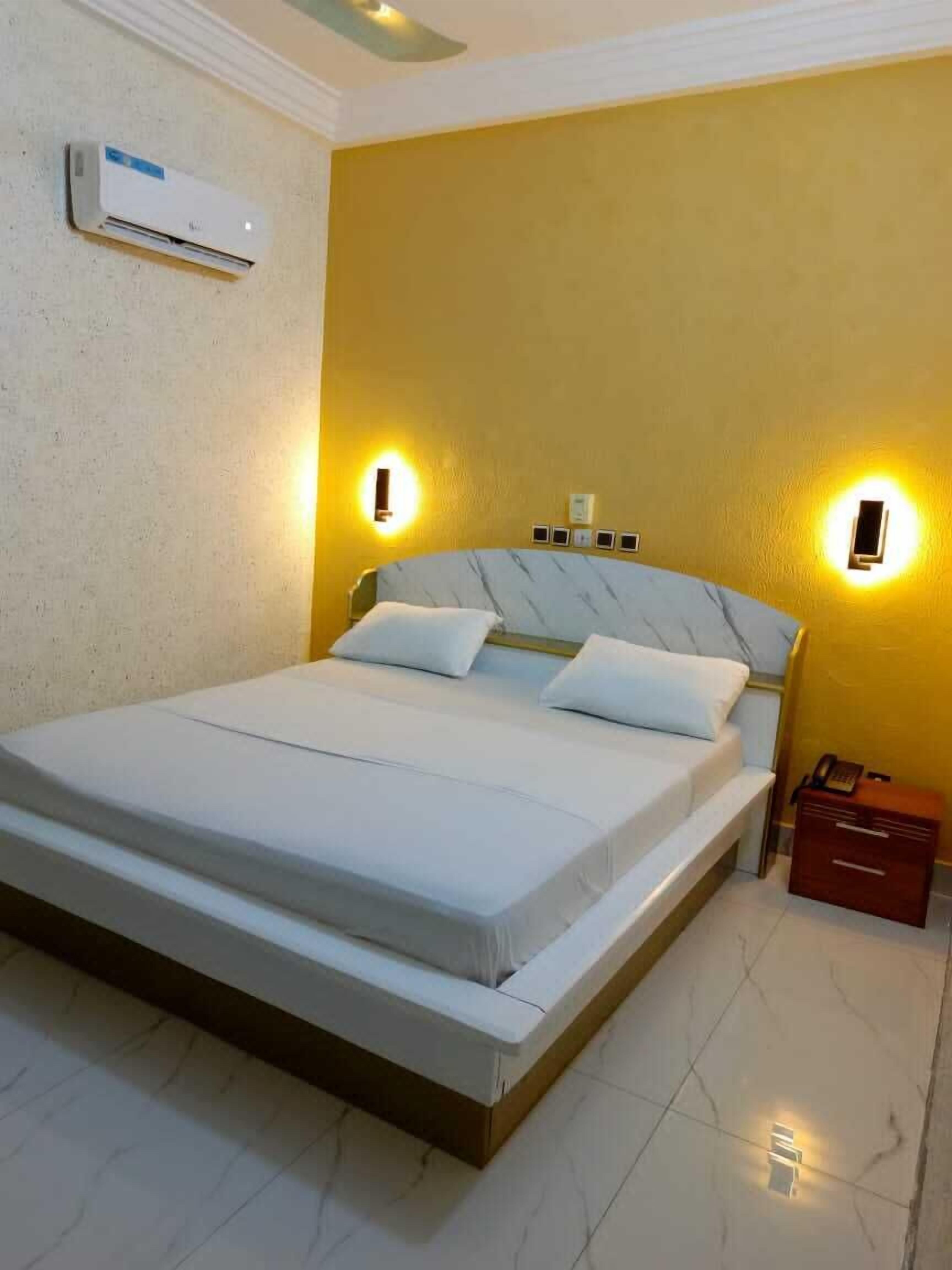 Una habitación de hotel con cama, mesitas de noche, aire acondicionado y luces en la pared.