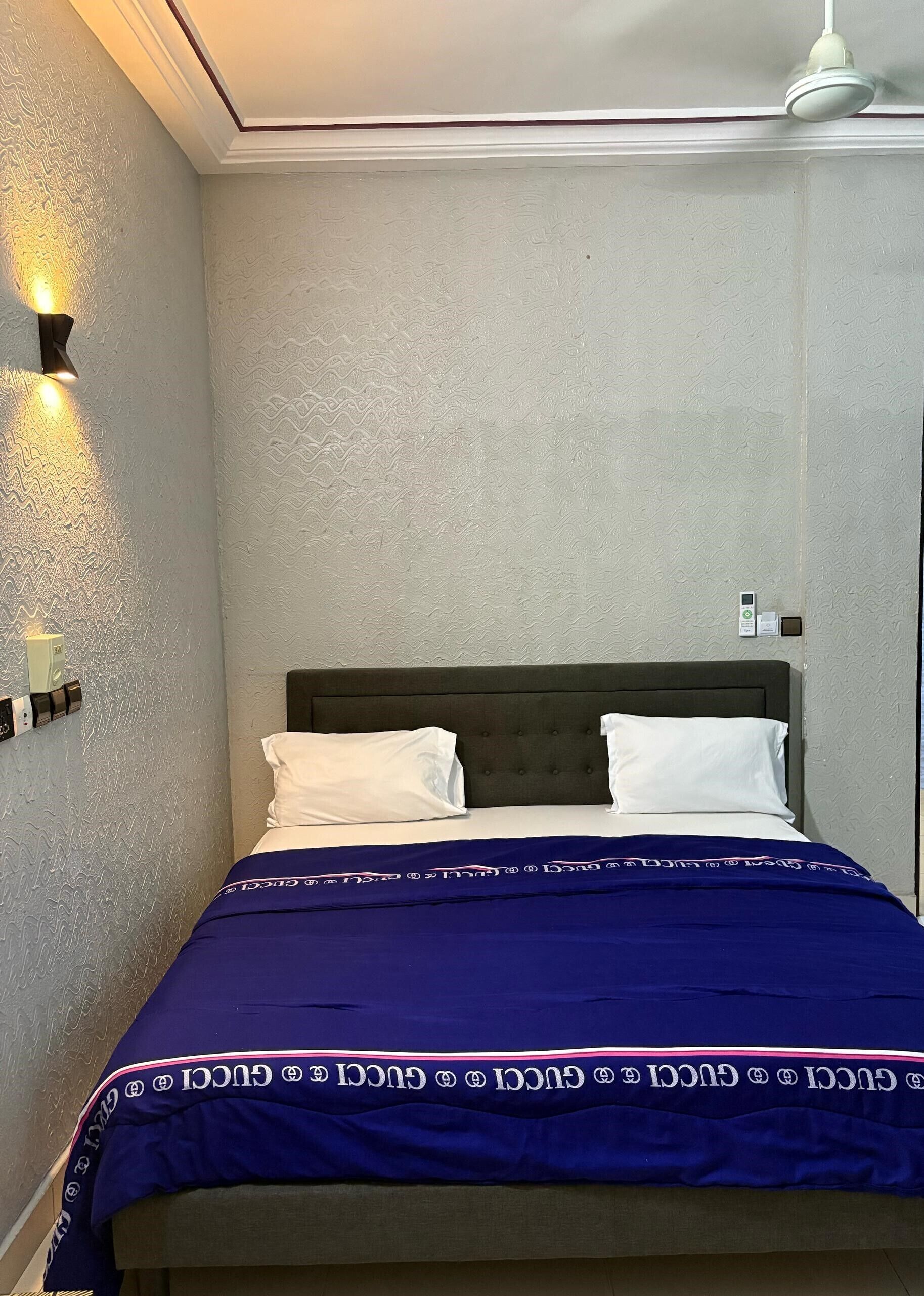 A bedroom with a bed, pillows, a blue blanket, a wall-mounted light, and a ceiling light.