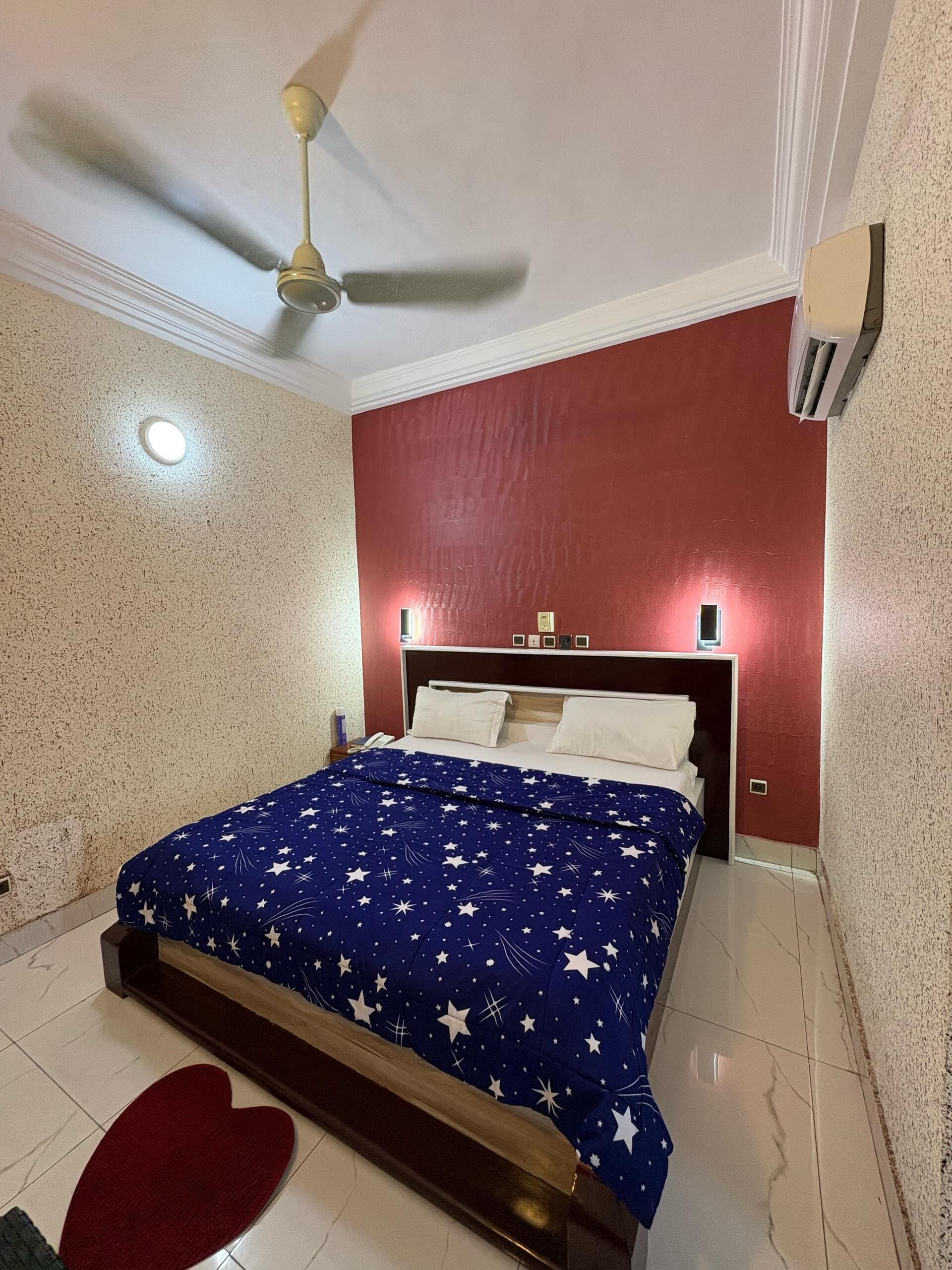Un dormitorio con una cama, ventilador de techo, mesita de noche y alfombra roja.