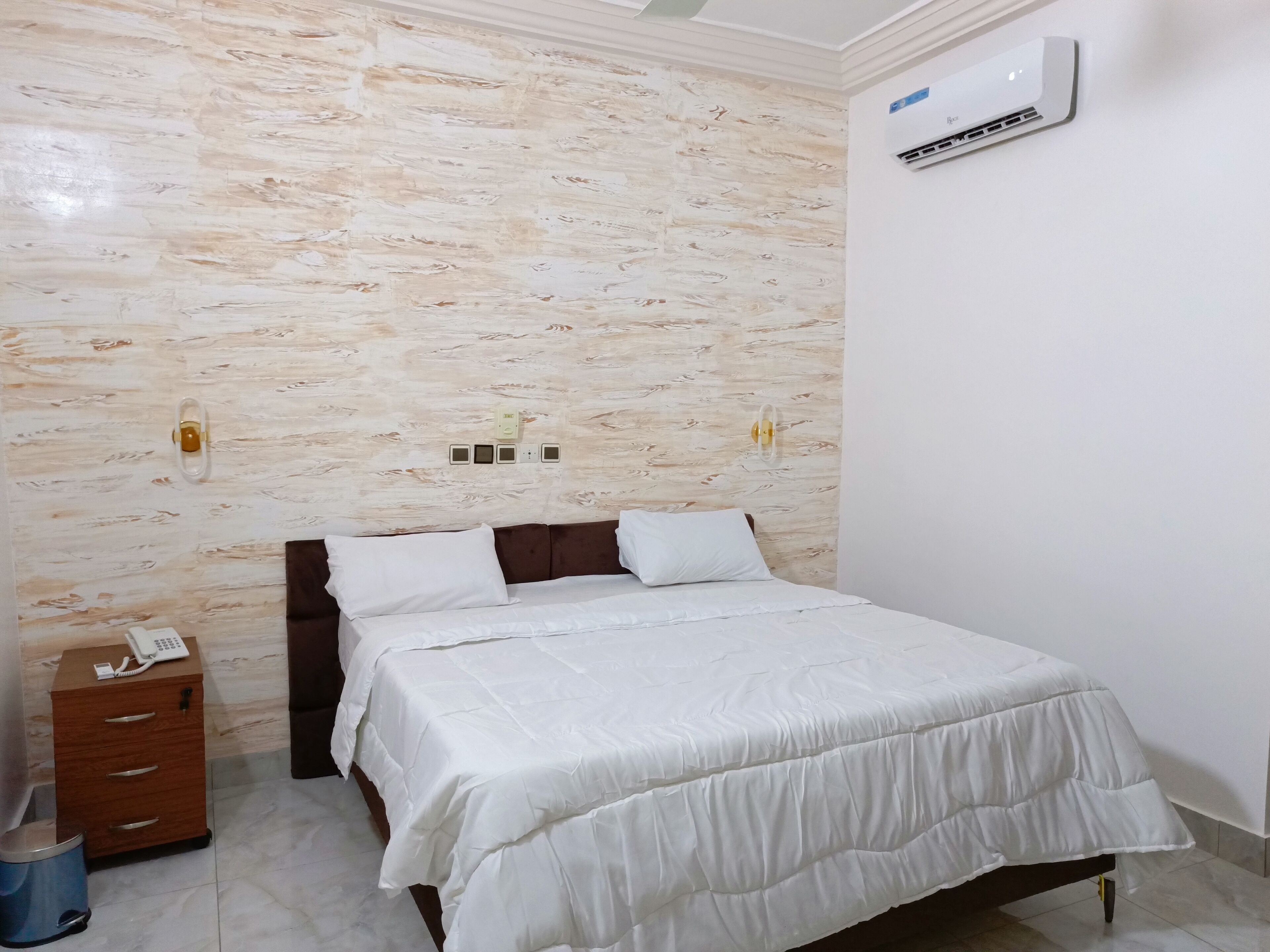 Una camera da letto con un letto, un comodino in legno, un telefono, un condizionatore d'aria a muro e una parete con motivo decorativo.