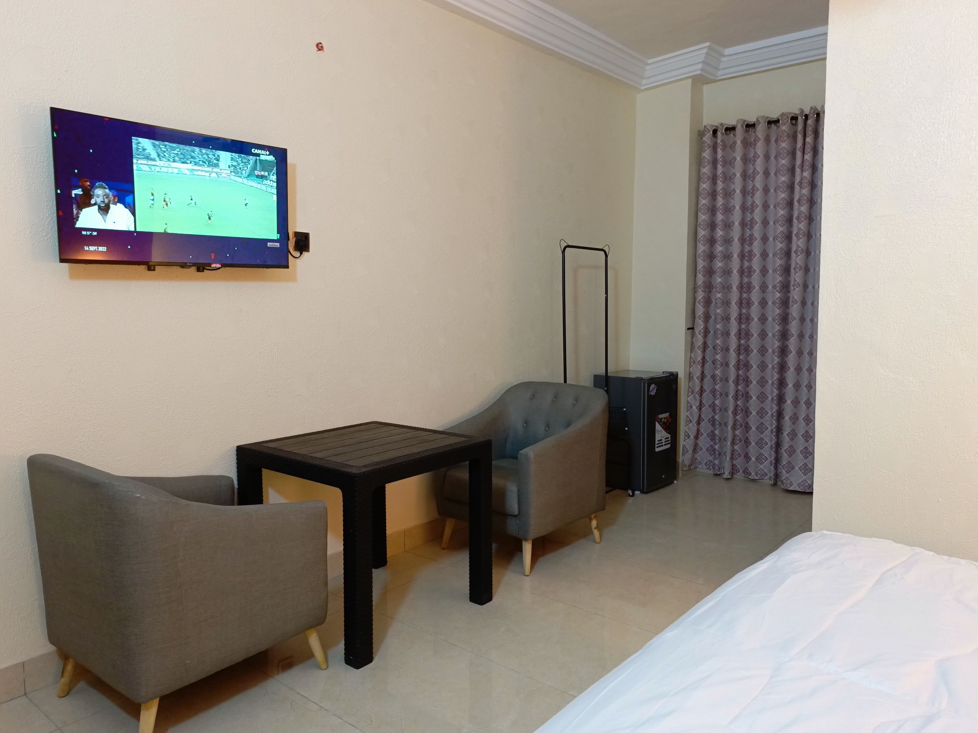 A hotel room with a bed, a TV mounted on the wall, a small table, two armchairs, and a floor lamp.